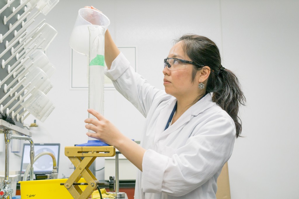 Lady in white lab coat pouring solution into beaker in lab